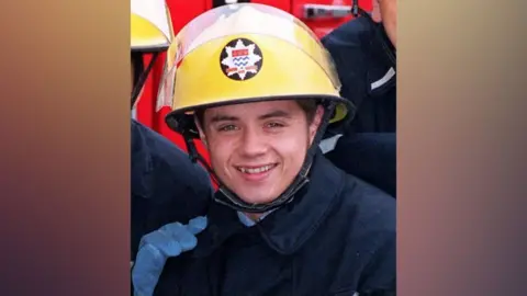PA Media Actor John Alford playing the part of a firefighter: He is wearing a yellow helmen, with a star-shaped badge, a blue coat and smiling.