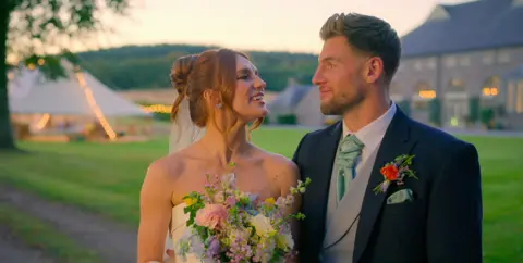 Netflix Megan and Kieran looking at each other smiling, wearing a wedding dress and suit respectively. A large countryside house can be seen in the background. 