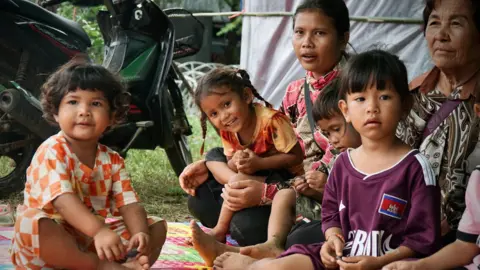 BBC/Lulu Luo Two women are sitting on the ground, with four children facing the camera, some of them smiling. Two of the little girls are on the women's laps. The other two are sitting on the floor and looking at the camera. Behind them is a motorcycle.