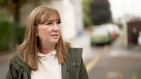 Lisa has long red hair and wears a green coat and beige top. She is standing by a road.