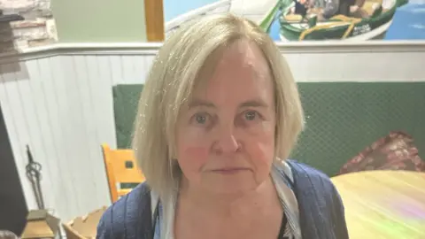 Bernadette McAliskey has shoulder length blonde hair and blue eyes. She is looking up at the camera at the picture as she stands next to a wooden table and chairs. She is wearing a blue cardigan and a white and blue blouse.