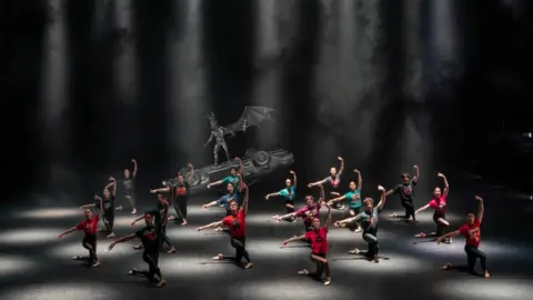 Johan Presson The dance company wearing black tights and colourful shirts on a lit black stage.