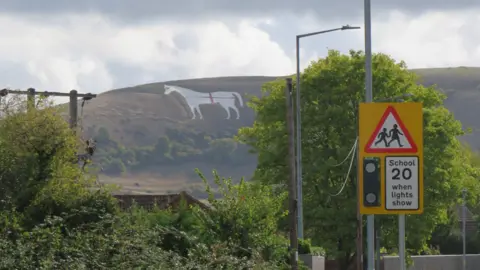 A large white horse carved into the side of a hill, with red fabric draped over it in a cross. In the foreground there are road signs and lampposts. 