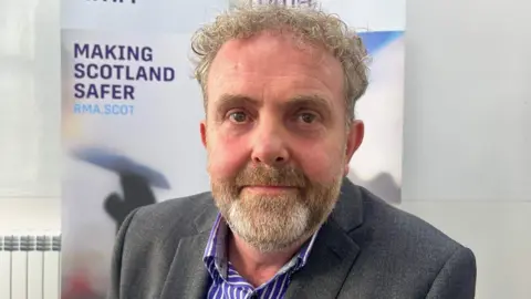 A man with a beard looks directly at the camera, wearing a grey suit and a striped shirt. A board behind him says Making Scotland Safer.