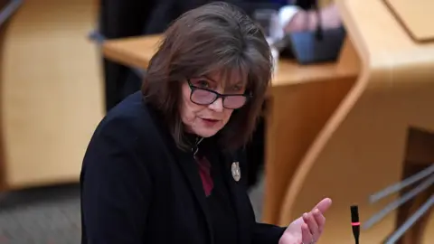 PA Media Jeane Freeman speaking in the Scottish Parliament in January 2021. She has shoulder length brown hair and glasses and is wearing a dark jacket with a brooch.