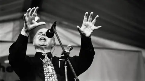 John Angerson A black and white image of the Jesus Army leader, Noel Stanton, delivering a sermon with his mouth open, eyes closed and hands grasping the air