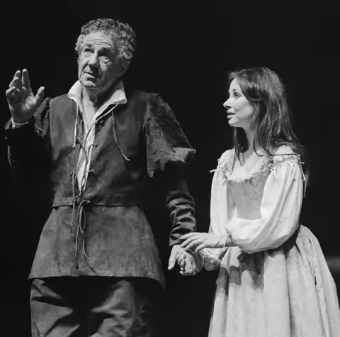 Joe Cocks Collection/Shakespeare Birthplace Trust Michael Hordern as Prospero  in rugged dark outfit with arm outstretched and the other being held by Miranda with brown hair and lovingly looking to Hordern in simple outfit