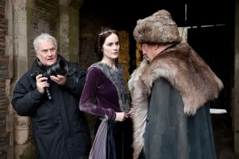 BBC/Neal Street Productions (L-R) Richard in dark blue anorak holding a camera, Michelle Dockery in purple velvet dress with grey fur in the centre, looking at Alun Armstrong (side profile) in brown fur hat and far round his shoulders and back and grey cloak