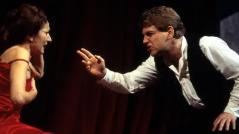 Reg Wilson/RSC Side profile of Jane Lapotaire with brown hair in a bun (playing Getrude) in red sleeveless dress with strap coming off her shoulder with an anguished expression being berated by Kenneth Branagh's Hamlet, dressed in white shirt and dark waistcoat -- who's gesticulating with one arm at Gertrude. 