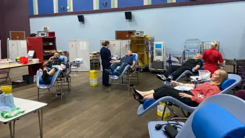 Ysgol Stanwell Four school staff members sit back in reclining chairs, with plasters and wires on their arms as they donate blood. Two nurses can be seen in the background preparing for the donations, along with empty vials on a nearby table.