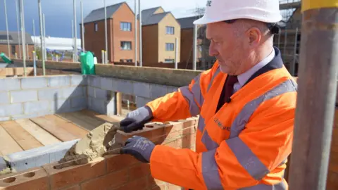 Housing Secretary Steve Reed, wearing a white hard hat and an orange high-vis suit, lays some bricks at a housing development near Houghton Regis in Bedfordshire.