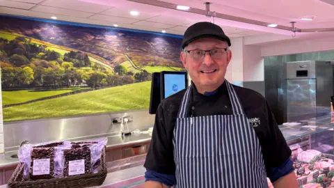 John Mettrick John Mettrick has a black flat cap and a blue and white striped butchers' apron on. He is wearing glasses and some grey hair is visible on the side of his head, under his cap. Behind him is a butchers' shop, with meats in cabinets and vacuum-packed goods in a basket on the counter.