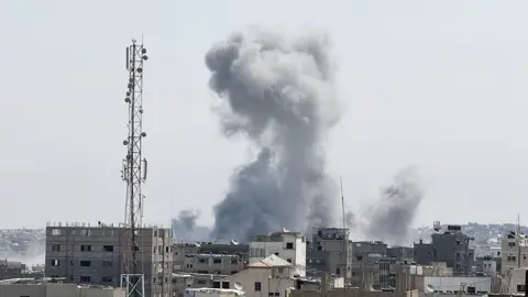 Anadolu via Getty Images A large plume of smoke rises into the sky from a densely built urban area, suggesting an explosion or fire. In the foreground, several multi-story buildings and a tall communication tower are visible on the left. The sky is clear, with no clouds apart from the smoke.
