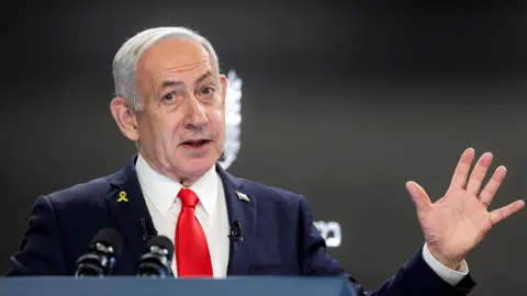 AFP via Getty Images Israeli Prime Minister Benjamin Netanyahu speaks at a podium with microphones in front of him. He wears a dark suit, white shirt, and red tie, with a yellow ribbon pin on his left lapel. His right hand is raised with fingers spread apart.