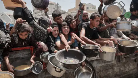 Anadolu via Getty Images A group of children reach over a barrier with various pots and pans, seeking food or water. The crowd includes men and women of different ages, with many arms extended in a chaotic scene. The cookware varies in size and type, including metal pots, plastic containers, and ladles. The background features the urban buildings and structures of Gaza City.