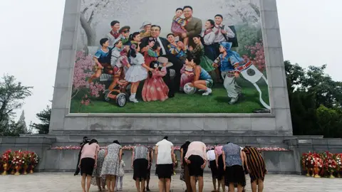 AFP via Getty Images A group of women standing in two-three rows bow in front of a mosaic in Pyongyang. The mosaic shows Kim Jong Un's father and grandfather smiling as they are surrounded by beaming children in a garden, flanked by pink flower bushes. On their the side of mosaic, which sits on a stone square, are large bouquets of red and white flowers in gold-coloured urns. The photo was taken on the 77th founding anniversary of the Kim family's regime.