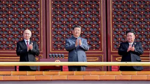 KCNA via Reuters (L-R) Russian President Vladimir Putin, Chinese leader Xi Jinping and North Korean leader Kim Jong Un standing in a line as they clap during a 
a military parade in Beijing on September 3, 2025. They are standing above the Gate of Heavenly Peace in Tiananmen Square. Behind them are tall, red ornate doors and a podium with mics. In front of them is the railing for the balcony they are on - it's gold-coloured with orange bricks.  