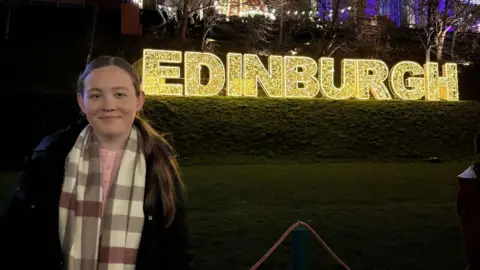 Handout Rebecca is standing on the left smiling at the photo. She is wearing a coat and a scarf. She is standing by the word Edinburgh which is lit up. The photo was taken at night. 