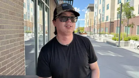BBC / Christal Hayes Scott Sperry is seen wearing a black T-shirt, a camo hat and sunglasses. He is outside in a courtyard of a student housing complex. 
