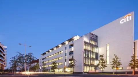 Donal McCann Citi's premises at the Gateway building in Belfast's Titanic Quarter. The large five-storey building has a grey facade, big windows and an illuminated "Citi" sign on the top right-hand corner.  It dwarfs a number of trees which line the street outside. 
