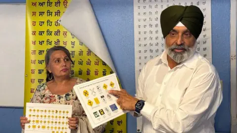 Gagandeep Singh An older man wearing a black turban and a white button-down shirt points to a sheet of paper with road symbols on it. Next to him is an older woman in a floral dress, holding a similar sheet with different symbols. They stand in front of two posters with Punjabi lettering