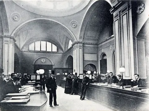 Corbis via Getty Images Black and white photo of one of the offices in the Bank of England in 1910 with men sitting behind wooden counters and others wearing top hats standing beside them
