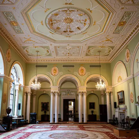 Bank of England Museum. Photo by Monica A Walker Vadillo The court room in the Bank of England which has decorated green walls, an elaborate gold, green, white and gold ceiling and a patterned rug with white columns and arches on the far side of the room