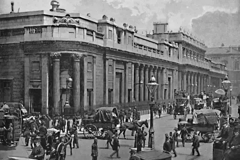 The Print Collector via Getty Images Black and white photo outside the Bank of England in 1896 with people walking on the street along with horse and carts and omnibuses 