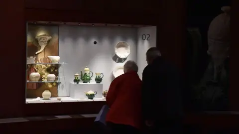 Getty Images A man and a woman are seen from behind looking inside the window of the Wedgwood museum. They are partially in shadow, the woman's red coat can be seen. Fine china, including teapots and vases, can be seen brightly lit on shelves. 