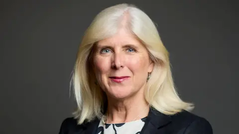 House of Commons A portrait image of a woman with long blonde hair and wearing a black jacket and white and black top. She is standing in front of a dark grey background.