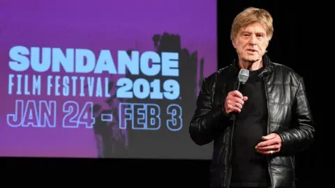 Getty Images Robert Redford is shown on stage at the Sundance Film Festival in 2019.  He is speaking into a microphone.