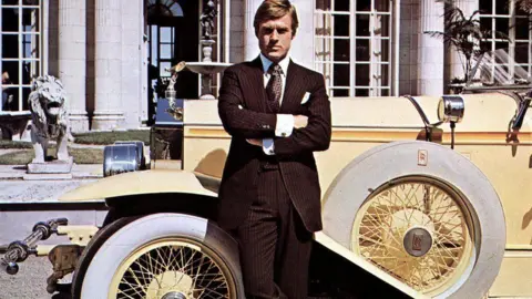 Getty Images Robert Redford is seen in a still photo promoting The Great Gatsby. He is standing in front of a vintage yellow Rolls Royce and has his arms crossed