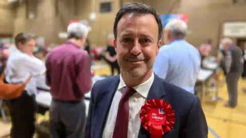 Simon Dedman, BBC Kevin Bonavia with short dark hair, smiling at the camera and wearing a blue jacket with a red Labour rosette, white shirt and red tie. He is at an election count and there are people standing around tables in the background.