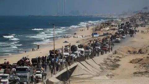 Reuters A long line of vehicles and trailers snake along a road, heavily packed with everything they can manage, as people flee south. The road is beachside, and you can see the ocean on the left, and in the distance is Gaza City.