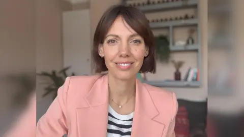 Menopause and Cancer Dani Billington is the founder of the charity Menopause and Cancer. She has brown hair and is wearing a light pink blazer and a black and white striped top. She is sitting in her living room. 