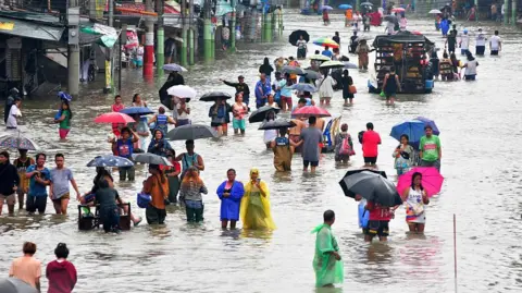 Getty Images Dozens of people holding umbrellas and wearing raincoats wade through a heavily flooded Manila street