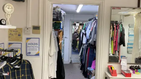 Indoor shot of the entrance to a charity shop clothing storeroom. In the foreground, on the left, a rack of hanging clothes holds a dark plaid button-up shirt. Next to it, a blue sign with yellow text reads "HOLDS Next Day Only" with a list of names underneath. A doorway in the center leads to a smaller room with more clothes hanging on racks. In the distance, a person is partially visible, walking away. On the right, a large mirror reflects the room and the clothing racks.