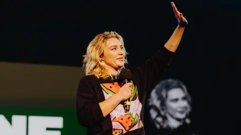 Luke Dyson Florence Pugh holds a microphone and waves. She wears a black jumper with a print showing slices of watermelon.