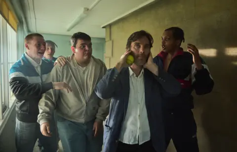 Netflix Cillian Murphy as headteacher Steve at the front of a group of boys who are all in various poses. Steve holds a tennis ball to his face and has his hands over his ears.
