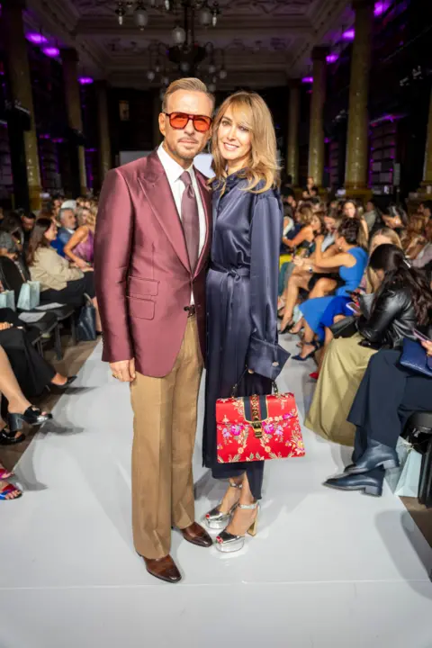 PA Media Matt Goss and his partner Chantal Brown on attending London Fashion Week. Goss is wearing a burgundy blazer with brown trousers and shoes. Brown is wearing a blue sleeved silk dress.