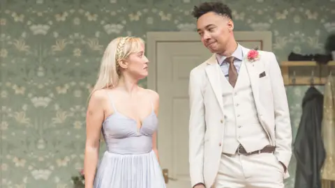 Mark Brenner Amelia Kenworthy and Kit Young star in Pickett's Emma. Kenworthy is wearing a light blue formal dress and Young is in a cream suit. They are mid-conversation.