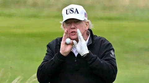 Getty Images President Trump wearing a white cap with USA on it and a black golf jacket. He has his hands to his mouth as he shouts. He is holding a golf ball in one hand and has a golf glove on the other