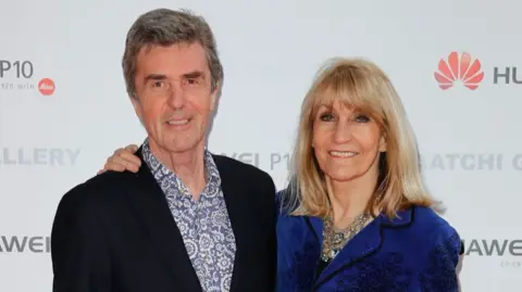 Getty Images John Stapleton and his wife  Lynn Faulds Wood arriving at the opening of an exhibition. Stapleton wears a black jacket with a floral shirt. Lynn Faulds Wood is wearing a blue jacket and large necklace. 