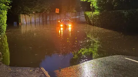 A car, with its headlights on, struggles through water in a flooded street