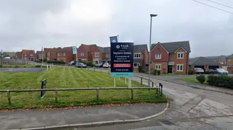 Google Google streetview of detached redbrick homes on the new estate, lining a road next to a green patch of grass.