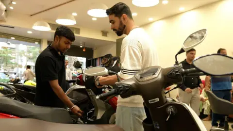 Vishnu Vardhan Two men - one in a black t-shirt and the other in white - looking at two-wheelers inside a showroom in Mumbai.