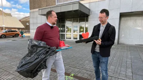 Two men standing outside a building. The man on the left is holding the Plook award and a bin bag while the man on the right is holding a crudely fashinoned arm model with a red heart jutting out of it
