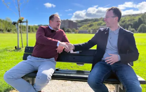 Two men sitting on a bench in a public park in the sun. They are shaking hands. 