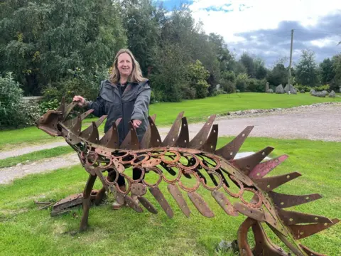 Woman stands behind a large rusted metal sculpture of some kind of reptile or dinosaur. It appears to have blunt spikes running along the length of its backed. At it's biggest, it is as tall as the woman's chest. 