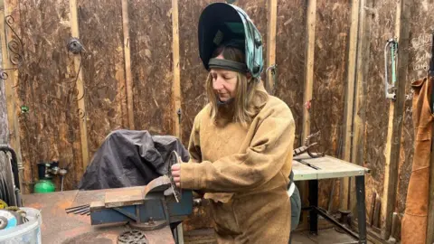 Woman in protective clothing standing next to a work bench. She has a large visor on her head which is raised. 